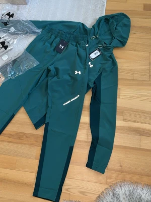 Grönt träningsset från Under Armour - Snyggt grönt träningsset från Under Armour med matchande hoodie och byxor. Setet har vita loggor, dragkedja på hoodien och elastisk midja på byxorna. Perfekt för träning eller chill. Materialet är mjukt och stretchigt för maximal komfort. Helt nytt 