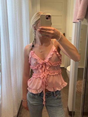 Rosa volang topp med knyt - Söt rosa linnetopp med volangdetaljer och knyt framtill. Toppen har djup v-ringning och tunna axelband, perfekt för sommarens varma dagar. Luftigt material och croppad passform som ger en trendig look.
