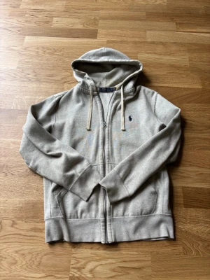 Beige hoodie från Polo Ralph Lauren - Snygg beige hoodie från Polo Ralph Lauren med dragkedja, huva med dragsko och klassisk blå logga broderad på bröstet. Tillverkad i mjukt bomullsmaterial och har två fickor framtill. Perfekt för en chill och stilren look.