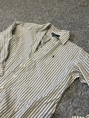 Randig skjorta från Ralph Lauren - Klassisk skjorta från Ralph Lauren i vitt och grått med vertikala ränder. Skjortan har lång ärm, knappar framtill och den ikoniska loggan broderad på bröstet. Tillverkad i bomull och har en avslappnad passform.