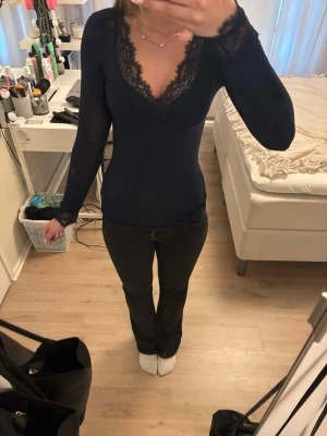 Mörkblå långärmad topp med spets - Snygg mörkblå långärmad topp med v-ringning och spetsdetaljer vid halsringningen och ärmsluten. Figurnära passform som ger en elegant look. Perfekt att styla med jeans eller kjol för en trendig outfit.