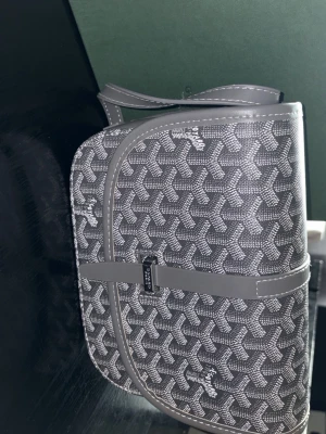 Grå mönstrad axelremsväska - Snygg grå axelremsväska med klassiskt grafiskt mönster och justerbar rem. Väskan har lock med magnetlås och detaljer i grått konstläder. Perfekt storlek för det viktigaste och ger en trendig touch till din outfit.