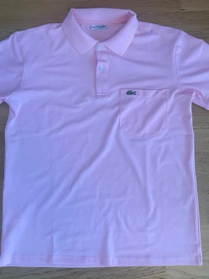 Ljusrosa pikétröja från Lacoste - Säljer en ljusrosa pikétröja från Lacoste med klassisk krage, knappar framtill och bröstficka med den ikoniska krokodilloggan. Perfekt till sommaren och enkel att matcha med jeans eller shorts.