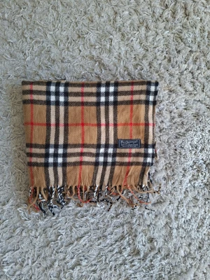 Rutig halsduk från Burberrys - Klassisk rutig halsduk från Burberrys i beige, svart, vitt och rött. Tillverkad i 100% lammull och har fransar i ändarna. Den ikoniska rutiga designen gör den superenkel att styla till olika outfits. Märkt 'Made in England'.