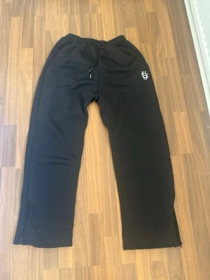 Suvene - black sweatpants - Svarta mjukisbyxor med avslappnad passform och resår i midjan samt dragsko. Byxorna har ett vitt tryck på vänster lår och dragkedjor vid bensluten för extra stil. Perfekta för chill dagar eller träning.