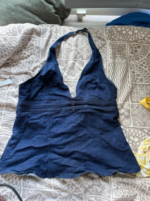 Mörkblå halterneck linnetopp - Snygg mörkblå linnetopp i halterneck-modell med djup v-ringning och dekorativa sömmar under bysten. Toppen har en figurnära passform och är tillverkad i ett denimliknande bomullsmaterial. Perfekt för dig som vill ha en trendig och unik topp till sommaren.