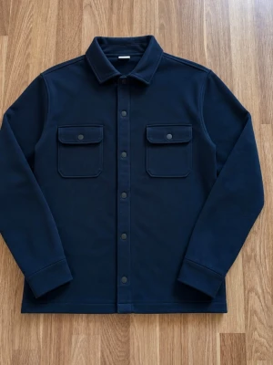 Mörkblå overshirt med fickor - Stilren mörkblå overshirt med klassisk krage och tryckknappar framtill. Två bröstfickor med lock och knappdetaljer ger en snygg look. Overshirten har raka ärmar och är tillverkad i ett mjukt, tjockare material som passar perfekt till lager på lager.