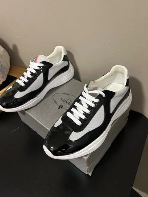 Svarta och vita sneakers med lackdetaljer från Zara - Snygga sneakers i svart och vitt med glansiga lackdetaljer och meshpaneler för extra andningsförmåga. Skorna har vita snören och en tjock, vit sula som ger en modern look. Perfekta för dig som vill ha en stilren och trendig sneaker till vardags.