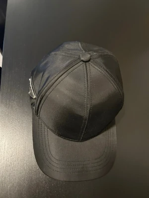 Svart klassisk keps - Snygg svart keps med böjd skärm och sex paneler. Kepsen har en enkel och stilren design med knapp på toppen och synliga sömmar. Tillverkad i ett glansigt tyg som ger en modern look. Perfekt accessoar för en avslappnad streetwear-stil.