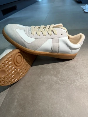 Vita sneakers med beige detaljer - Stilrena sneakers i vitt med beige mockadetaljer och beige snörning. Skorna har en klassisk låg siluett och en gummisula i ljusbrunt med mönstrad undersida. Materialmix av läder och mocka ger en snygg kontrast och modern look.