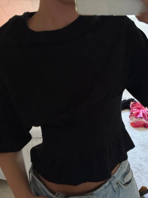Svart topp med markerad midja - Snygg svart topp med korta ärmar och markerad midja. Toppen har en lätt volang nertill och rund halsringning. Perfekt till jeans eller kjol för en trendig look.