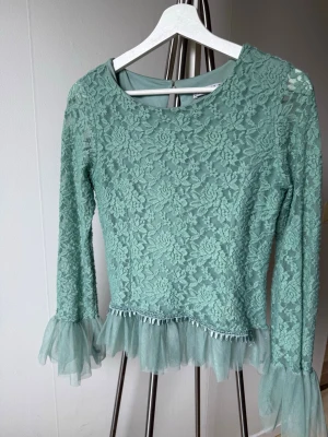 Grön spetsblus med volangdetaljer - Snygg grön blus i spets med långärmad design. Blusen har volangdetaljer i tyll vid ärmslut och nederkant samt ett blommigt spetsmönster över hela plagget. Perfekt för dig som vill ha en romantisk och trendig look.
