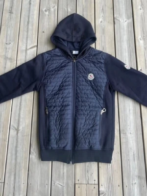 Mörkblå Moncler hoodie med dragkedja - Snygg mörkblå hoodie från Moncler med quiltad front och två dragkedjeförsedda fickor. Klassisk Moncler-logga på bröstet och ärmen. Huva och muddar vid ärmslut och nederkant. Perfekt för en chill och stilren look.