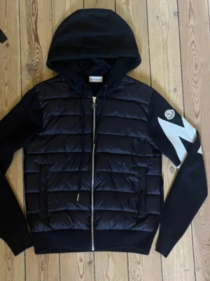 Moncler  - Svart cardiganjacka från Moncler med vadderad front, stickade ärmar och stor huva. Ena ärmen har ett vitt zigzag-mönster och Moncler-logga. Jackan har dragkedja framtill och två fickor med dragkedja. Perfekt för en trendig och sportig look.