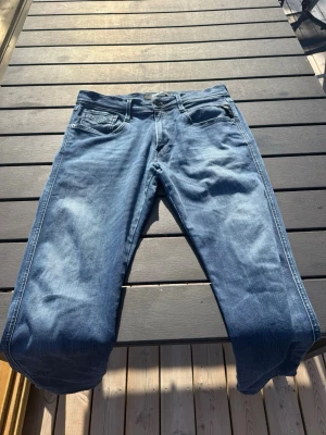 Replay anbass  - Säljer ett par klassiska blå jeans från Replay i rak modell. Jeansen har fem fickor, snygga slitningar och ljusare tvättade partier framtill. Tillverkade i denim med detaljerade sömmar och Replay-logga på fickan. Perfekta för en avslappnad och trendig look.