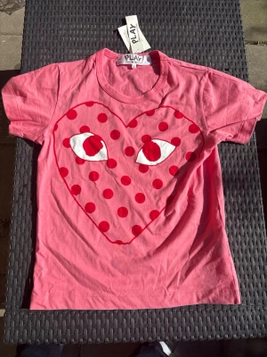 Rosa t-shirt med hjärta och prickar - Cool rosa t-shirt med ett stort rött hjärta med ögon och prickar på bröstet. T-shirten har rund hals och korta ärmar, perfekt för dig som vill sticka ut. Materialet känns mjukt och skönt, troligen bomull.
