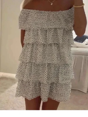 Vit offshoulder volangklänning - Söt vit kortklänning med svarta prickar och flera volanglager. Offshoulder-modell som ger en trendig look. Klänningen har en svart knyt i midjan som markerar siluetten. Perfekt för sommarens alla tillfällen.