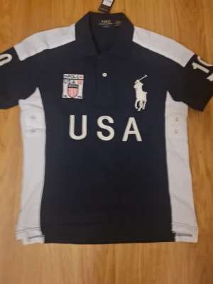 Mörkblå Polo Ralph Lauren piké - Mörkblå pikétröja med vita detaljer från Polo Ralph Lauren. Stora broderade bokstäver 'USA' på bröstet, polospelare-logga och patch med amerikansk flagga. Klassisk krage och knappslå framtill, samt vita paneler på sidorna och ärmarna.