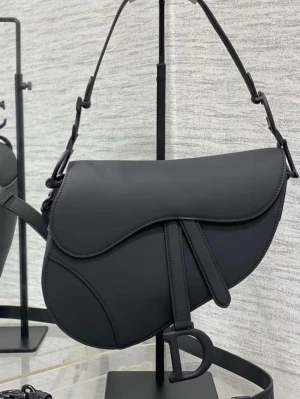 Dior Väska - Stilren svart handväska i sadelmodell med unik form och justerbart axelband. Väskan har dekorativa sömmar och ett stort D-format hänge framtill. Tillverkad i slätt läder som ger en lyxig känsla och passar perfekt till trendiga outfits.