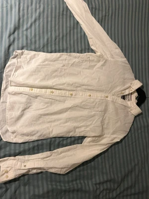 Vit långärmad skjorta i bomull - Klassisk vit skjorta med lång ärm och knappar framtill. Skjortan har en enkel krage och bröstficka, tillverkad i mjuk bomull. Perfekt för en clean och stilren look.