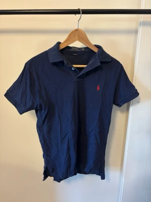 Marinblå pikétröja från Polo Ralph Lauren - Klassisk marinblå pikétröja från Polo Ralph Lauren med korta ärmar och röd broderad logga på bröstet. Tröjan har krage och knäppning framtill. Perfekt för en stilren och avslappnad look.