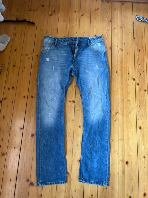 Blå raka jeans med slitningar - Säljer ett par blå jeans med rak passform och snygga slitningar framtill. Jeansen har klassisk femficksdesign och ljusare partier på låren och knäna för en cool, avslappnad look. Perfekta till vardagsstilen och gjorda i denim. Köpta för länge sedan men använda men i fint skick. 