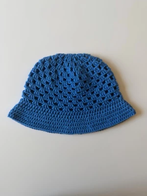 Blå virkad bucket hat - Handgjord bucket hat i virkad bomull, i en härlig blå nyans. Hatten har ett luftigt mönster och brett brätte, perfekta för en trendig och avslappnad stil. Säljs styckvis. 