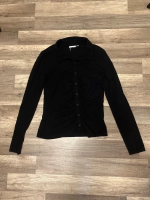 Svart elegant rynkad topp med knappar – Strl M | Använd 1 gång | Nyskick  - 🖤 Superfin svart topp i storlek M 🖤  Så fin och stilren topp som passar till precis allt! Den smickrande rynkade designen framtill ger en feminin och elegant look, samtidigt som den är bekväm att bära. Perfekt både till vardags, jobbet eller en utekväll.  ✨ Endast använd en gång, därför är den i mycket fint skick – ser nästan ny ut! Ett tidlöst plagg som är lätt att matcha med jeans, kjol eller kostymbyxor.