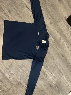 Mörkblå Nike PSG träningströja - Säljer en mörkblå långärmad träningströja från Nike med PSG-logga på bröstet och vit Nike Swoosh. Tröjan har halv dragkedja och är tillverkad i Dri-FIT-material som andas, perfekt för träning eller sportaktiviteter.