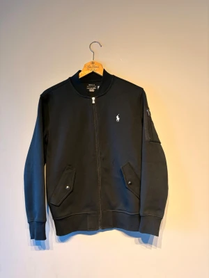 Svart bomberjacka från Polo Ralph Lauren - Stilren svart bomberjacka från Polo Ralph Lauren med klassisk broderad logga på bröstet. Jackan har ribbade muddar, dragkedja framtill, fickor med tryckknappar och en ärmficka. Tillverkad i mjukt bomullsmaterial för en bekväm känsla.