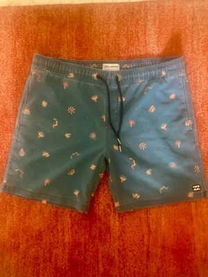 Billabong badshorts - Snygga badshorts från Billabong i en fin blå nyans med diskret tropiskt mönster. Perfekta för stranden, poolen eller semestern.  * Märke: Billabong * Färg: Blå med flerfärgat mönster * Storlek: M. Jag har 32 i midjan och de är för stora för mig. Uppskattar storleken till 33/34. * Skick: mycket bra, inga synliga hål eller fläckar. * Resår och dragsko i midjan för bra passform. * Sidofickor.  MÅTT Midja: 48cm (mätt liggandes på golvet). Benlängd: 43cm (yttersöm). Gren: 29cm  Spana gärna in mina öv