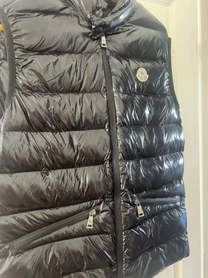 Svart Moncler väst  - Snygg svart dunväst från Moncler med glansig finish och klassisk logga på bröstet. Västen har hög krage, dragkedja framtill och två fickor med dragkedja. Perfekt för lager-på-lager och streetstyle.