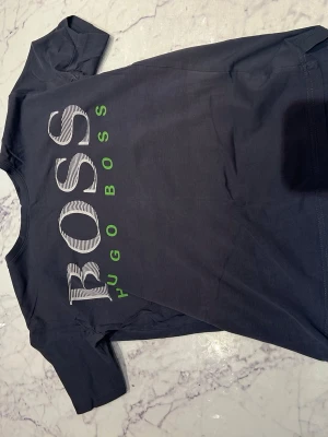 Svart Hugo Boss t-shirt med logga - Snygg svart t-shirt från Hugo Boss med stor vit och grön logga på framsidan. Klassisk passform och rund halsringning. Perfekt till jeans eller shorts för en clean och stilren look.