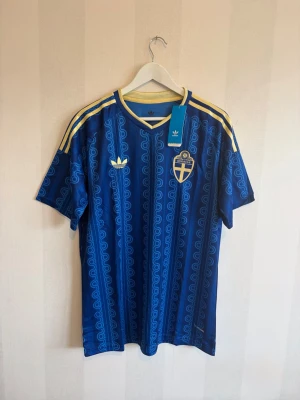 Blå Sverige fotbollströja Adidas - Säljer en blå fotbollströja från Adidas med Sveriges landslagsemblem på bröstet. Tröjan har gula detaljer, tre ränder på axlarna och ett unikt mönster i olika blå nyanser. Perfekt för match eller träning. Materialet är lätt och andas.