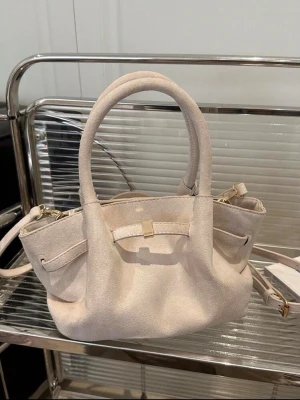 Beige mocka handväska med dubbla handtag - Stilren beige handväska i mjuk mocka med två rundade handtag och avtagbar axelrem. Väskan har en enkel design med dekorativ rem och guldfärgad detalj framtill. Perfekt storlek för det viktigaste och ger en lyxig känsla till din outfit.