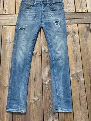 Blå slitna jeans med raka ben - Säljer ett par blå dondups med raka ben och slitningar på låren och knäna. Jeansen har en klassisk femficksmodell, ljus tvätt och detaljer som små färgstänk för en trendig look. 