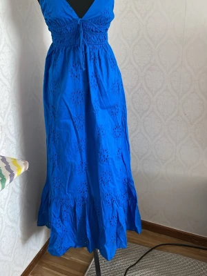 Blå långklänning med broderade detaljer - Snygg blå långklänning med v-ringning och smock i midjan. Klänningen har broderade hålmönster och volang längst ner. Perfekt för sommarens alla tillfällen och har en feminin siluett.