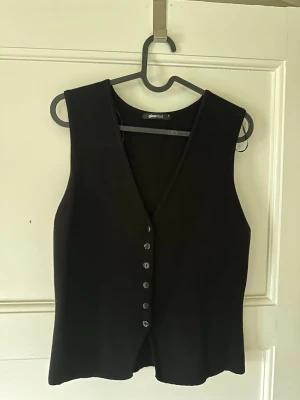 Svart väst med knappar - Stilren svart väst från Gina Tricot med djup V-ringning och fem knappar framtill. Västen har en rak passform och är tillverkad i ett mjukt tyg som ger en clean look. Perfekt att styla över en skjorta eller t-shirt.