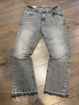 Gråa jeans från ZARA - Säljer ett par gråa raka jeans från ZARA med slitna och fransiga benslut. Jeansen har klassisk femficksdesign och är tillverkade i denim. Perfekta för en avslappnad och trendig look.