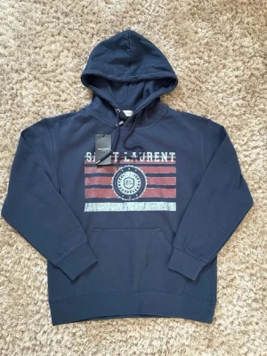 Mörkblå hoodie från Saint Laurent - Snygg mörkblå hoodie från Saint Laurent med tryck i vitt och rött framtill. Klassisk huva med dragsko och känguruficka. Tillverkad i mjuk bomull för skön känsla. Perfekt för dig som vill ha en stilren och trendig look. Storlek L men passar M (saknar tagg nu och endast testad)