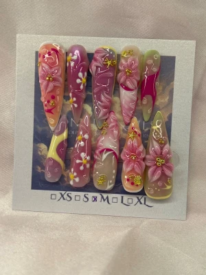 LuxeFantasy (M);Extra Long Stiletto - Set handgjorda unika press-on naglar i storlek M: 16 mm, 12 mm, 12,5 mm, 11,5 mm, 10 mm med en extra lång, stiletto tippar. Naglarna har en mix av rosa, nude, lila och gul bas med handgjorda 3D-blommor, små vita blommor, guldglitter och detaljer i vitt. 