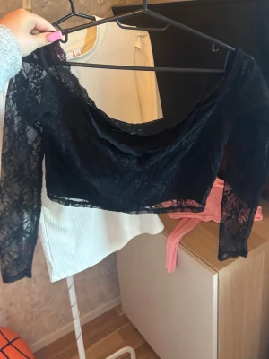 Svart croppad spetsblus - Säljer en svart croppad blus i spets med långärmad design. Toppen har en snygg offshoulder-skärning och transparent spetsmaterial som ger en trendig look. Perfekt för dig som vill sticka ut lite extra.