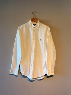 Vit skjorta från Polo Ralph Lauren - Klassisk vit skjorta från Polo Ralph Lauren med broderad logga på bröstet. Skjortan har lång ärm, knappar framtill och krage. Perfekt till både jeans och chinos för en clean och stilren look.