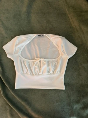 Vit croppad topp PrettyLittleThing - Säljer en vit croppad topp från PrettyLittleThing med korta ärmar. Toppen har en söt knytning framtill och är gjord i ett mjukt, stretchigt material. Det finns en liten dammfläck vid den nedre delen som uppstått i samband med flytt men tröjan kommer att fraktas nytvättad! 