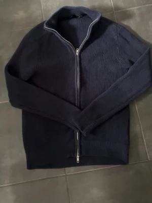 Mörkblå ribbstickad ziptröja - Mörkblå ribbstickad tröja med dragkedja hela vägen framtill och hög krage. Tröjan har långa ärmar och en stilren, enkel design som passar till mycket. Perfekt för lager på lager och håller dig varm under kyliga dagar.