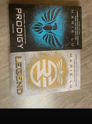 Legend & Prodigy av Marie Lu - Två dystopiska ungdomsromaner av Marie Lu: 'Legend' med vit och guld omslag samt 'Prodigy' med svart och blått omslag. Båda böckerna har moderna, grafiska omslag och är utgivna av Modernista. Perfekt för dig som gillar spänning och framtidsvärldar.