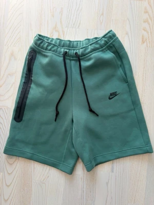 Gröna Nike tech shorts  - Snygga gröna shorts från Nike med svart snörning i midjan och en dragkedjeförsedd ficka på sidan. Tillverkade i mjukt material som passar perfekt för avslappnade dagar. Har endast provats men lapparna har klippts av tyvärr. 