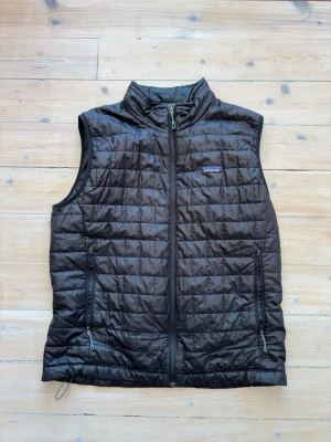 Svart dunväst från Patagonia - Snygg svart dunväst från Patagonia med quiltat mönster och dragkedja framtill. Västen har två dragkedjeförsedda fickor och hög krage. Perfekt för lager-på-lager och outdoor-vibe. Tillverkad i lätt och värmande material.