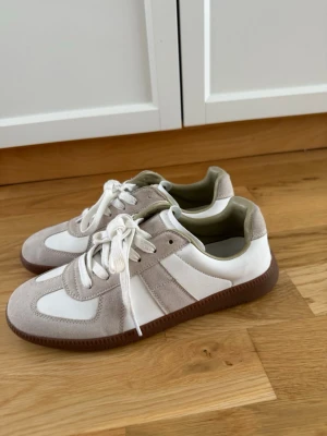 Vita GATS sneakers  - Snygga sneakers i vitt och beige med ovandel i mocka och släta partier i storlek 41. Skorna har vita snören och en brun gummisula som ger en retrovibe. Perfekta för dig som gillar stilrena och klassiska sneakers med en twist.