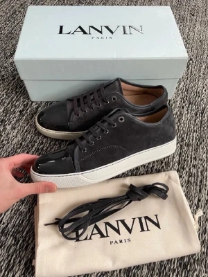 Svarta Lanvin sneakers i mocka och lack - Snygga svarta sneakers från Lanvin med ovandel i mocka och tåparti i blankt lack. Skorna har vita sulor och svarta skosnören. Kommer med originalkartong och dustbag. Perfekta för dig som gillar stilrena och exklusiva sneakers.